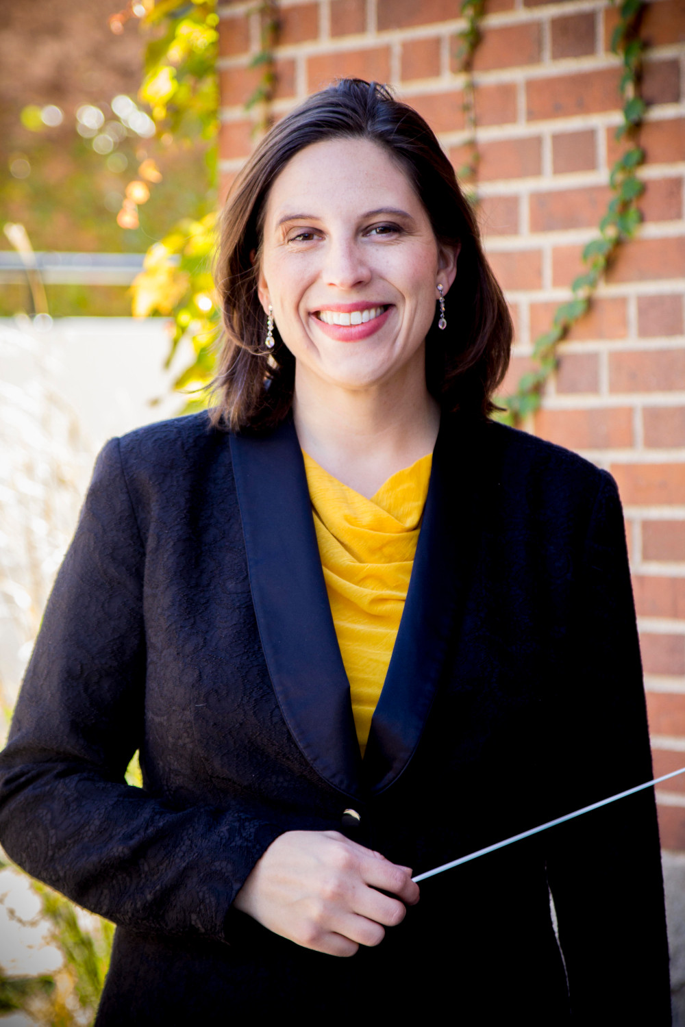 Image shows Seward Concert Band Director, Cassandra Bechard. Bechard is a white female wearing a dark suit and holding a baton.